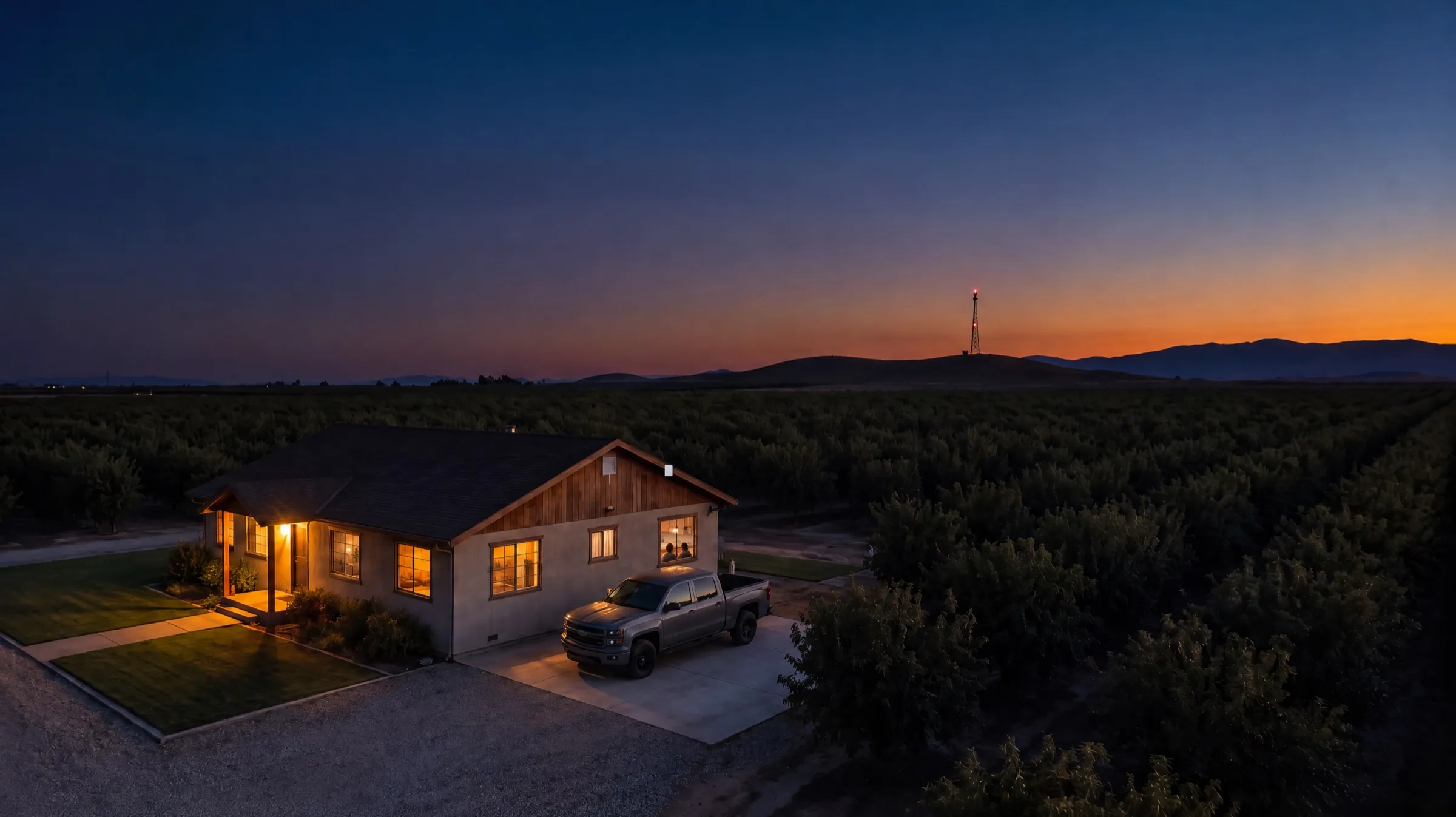 A farmhouse at dusk with a gray pickup in the driveway, a small radio on the eave and a tower on the hills behind it