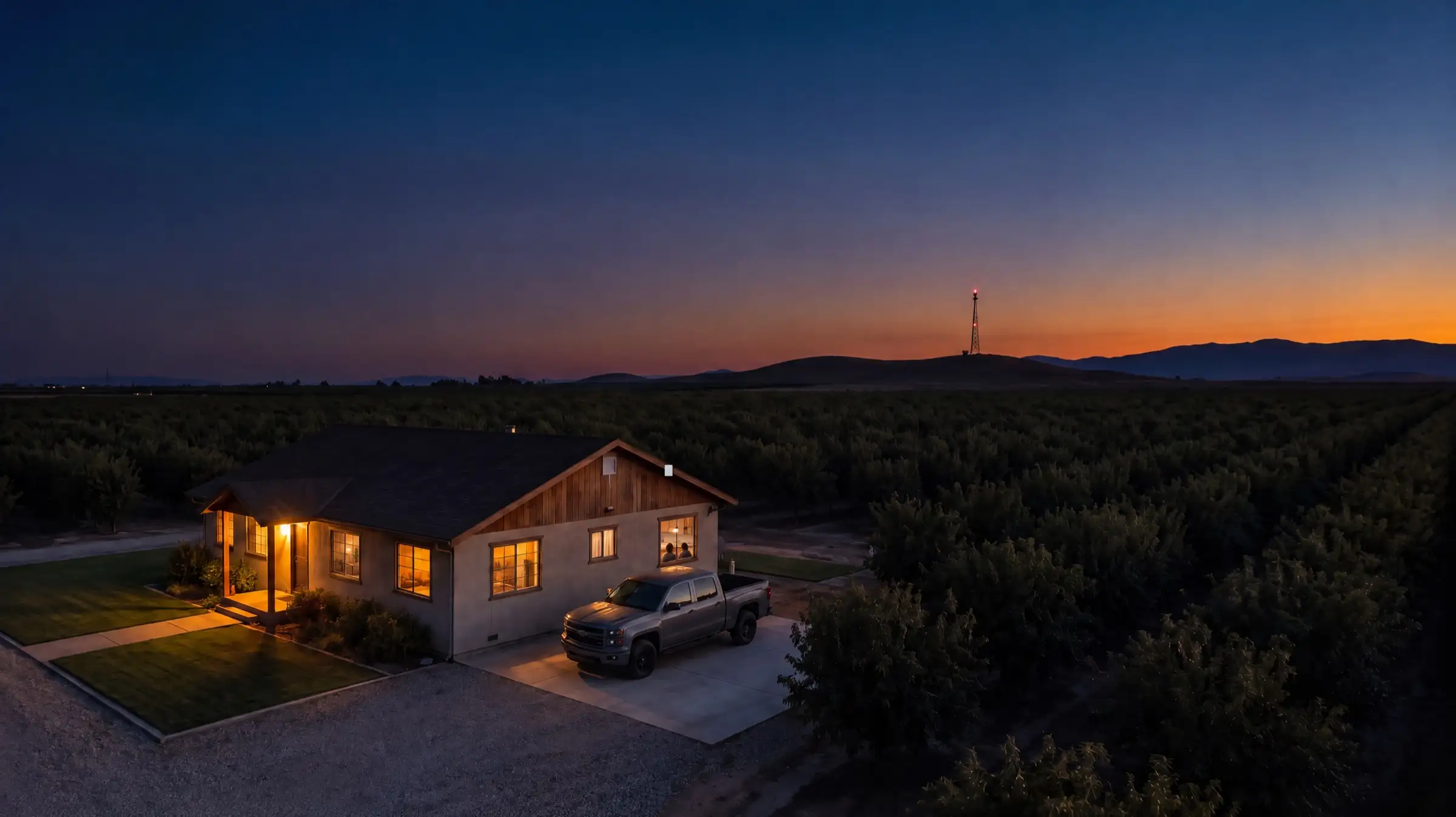 Una casa de rancho al atardecer con una camioneta gris en la entrada, un radio pequeño en el alero y una torre en las colinas detrás