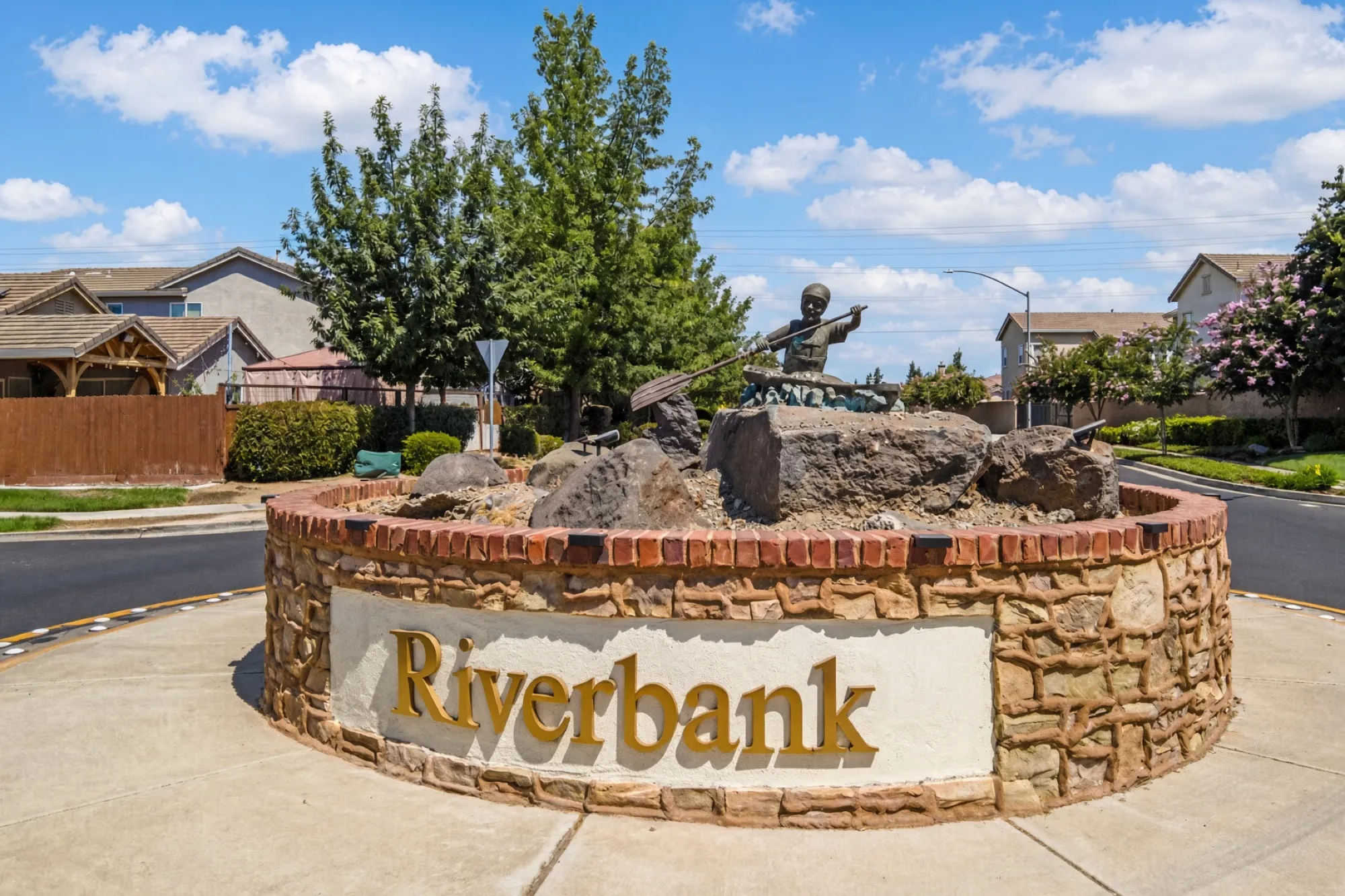 A stone Riverbank welcome sign with a statue of a rafter in a neighborhood roundabout