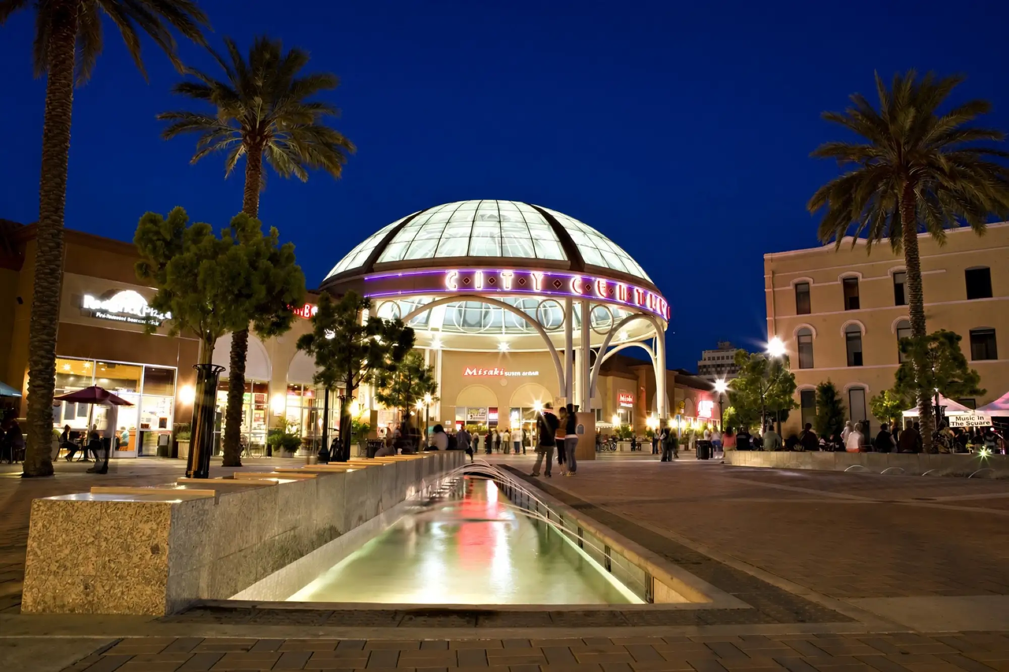 Una cúpula de vidrio iluminada sobre una plaza con palmeras en el centro de Stockton de noche